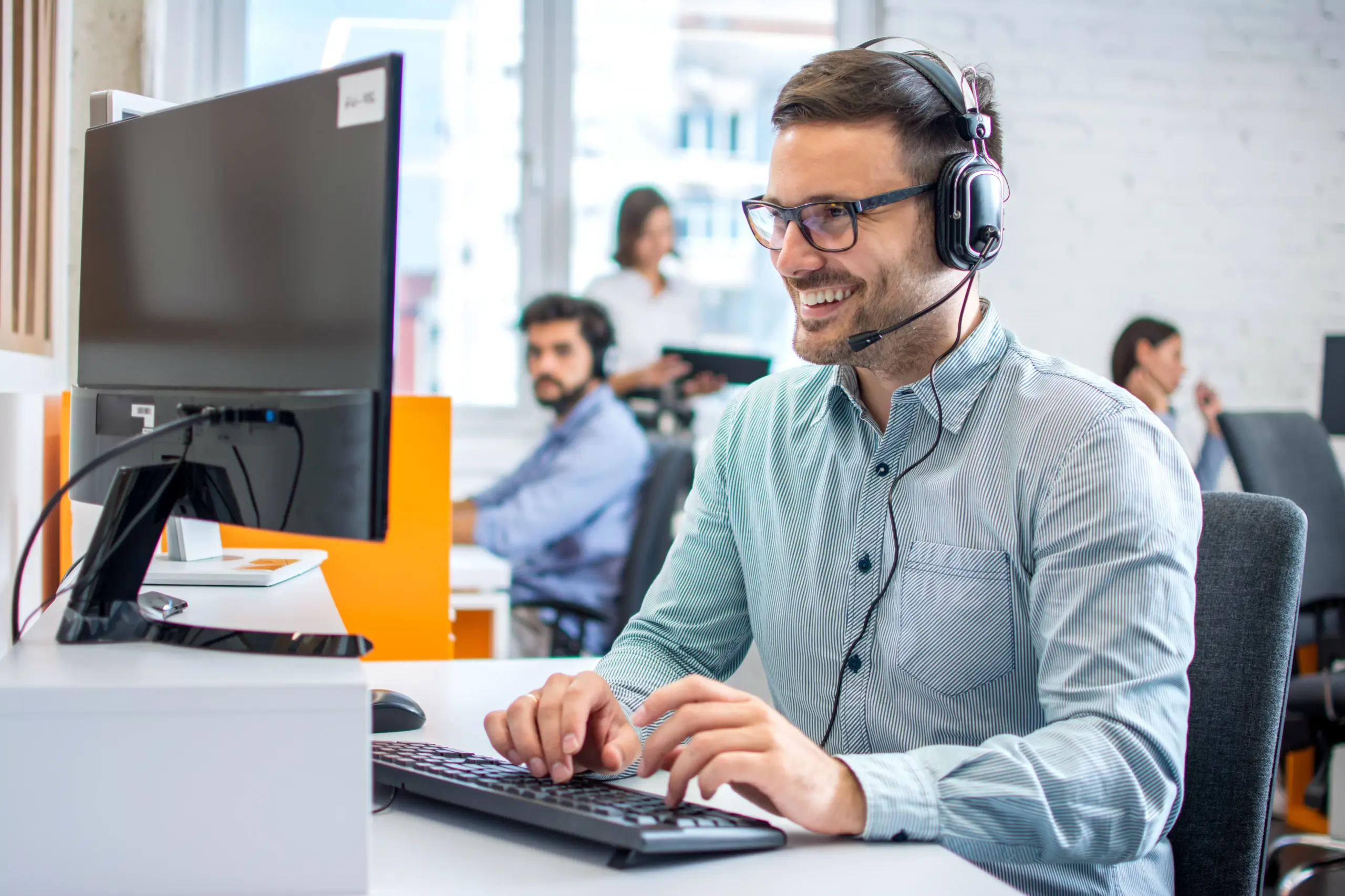 A stock image of a man smiling while working at a computer. Others are working in the background.